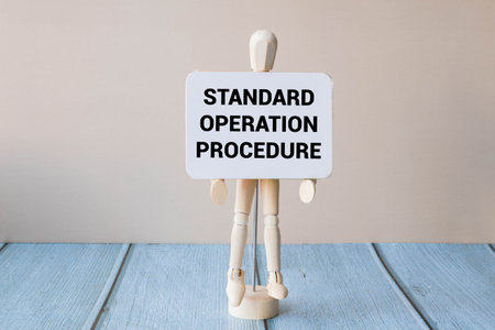 The image shows a wooden figurine holding a sign with the text Standard operation procedure on a blue wooden surface. The background is plain and neutral.の写真素材