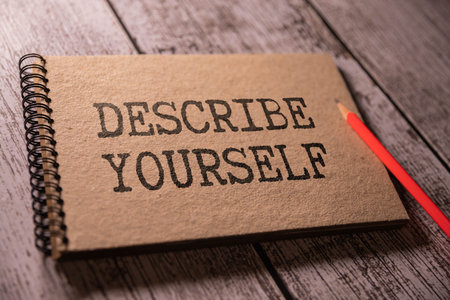 A red pencil is on a brown notebook with black text reading Describe Yourself. The notebook is on a wooden background.の写真素材