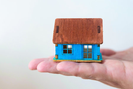 Wooden house model in male hand on background of new buildings. Real estate agent, choice between a country cottage or a city condoの写真素材