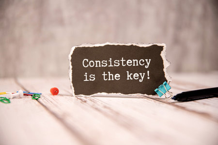 A piece of paper with the handwritten message Consistency is the key is taped to a wood grain surface. The paper has a torn edge.の写真素材