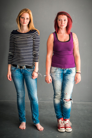 Two angry women connected by a pair of handcuffs, blond and red-haired, wearing jeans, barefoot and red chucks on grey backgroundの写真素材