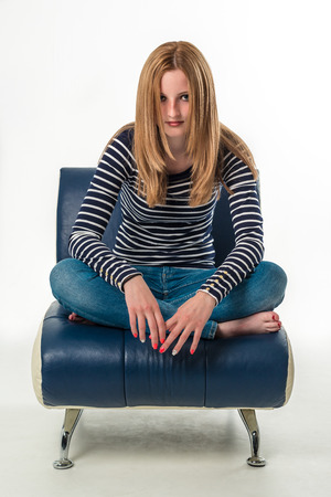 Beautiful stylish blonde young woman sitting cross-legged on a small blue couch over white backgroundの写真素材