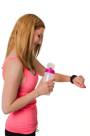Young blonde woman in pink shirt looking at digital watch with bottle in her hand, isolated on whiteの写真素材