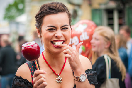 Attractive young woman in traditional Dirndl dress holding a red caramelized apple at the Oktoberfest with people in the background.の写真素材