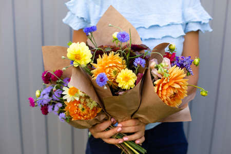 Woman in blue blouse holding beautiful bouquets of flowers. Hands with bouquets of seasonal flowers in craft paper. Bouquets as a gift on 1st september. Orange dahlia flowersの写真素材