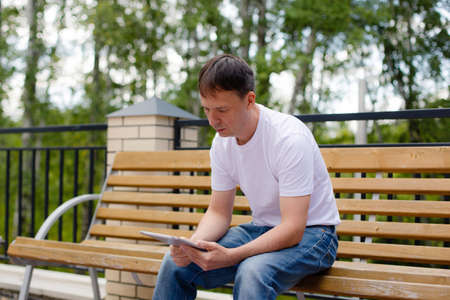 Portrait of a man in the park holding tablet and sitting on a bench. Man in white t-shirt and blue jeans with tablet.の写真素材