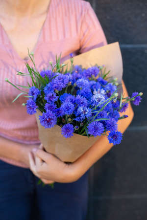 Woman in pink t-shirt holding a bouquet of blue cornflowers. Bouquet of blue centourea in craft paper in the hands. Grey dark backgroundの写真素材