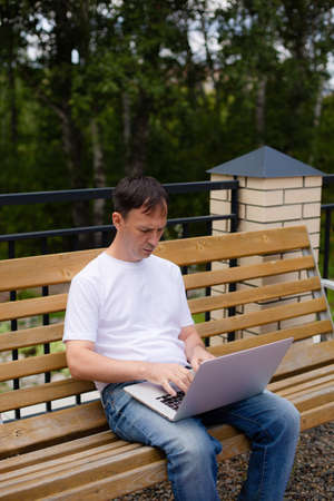 Portrait of a middle-aged man with laptop sitting on the bench. Man in white t-shirt and blue jeans with laptop sitting on the bench in the park.の写真素材