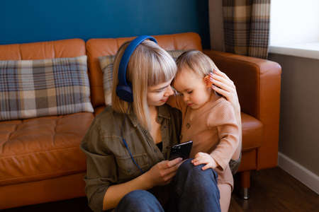 Young woman with headphones and baby at home. Mother with one year old child listening to music at home. Woman with phone, headphones and child sitting on the floor at home.の写真素材