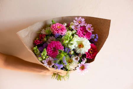 Hand with beautiful bouquet of seasonal flowers on pink background. Pink zinnia, white dahlia, cosmos, purple ageratum, white antirrhineae. Bouquet of seasonal flowers in craft paper.の写真素材