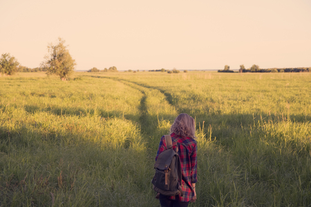 girl in plaid shirt with a backpack on shoulders walking down country road in the middle of a field with tall grassの写真素材