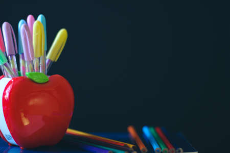School accessories. Multicolored Pencils Stand at the stands in the shape of an apple. In Background of a dark school board and a piece of chalk. On the board is a place for a copy space.の写真素材