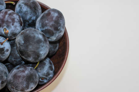 Several ripe blue plums in a deep clay plate on a white background with a copy spaceの写真素材