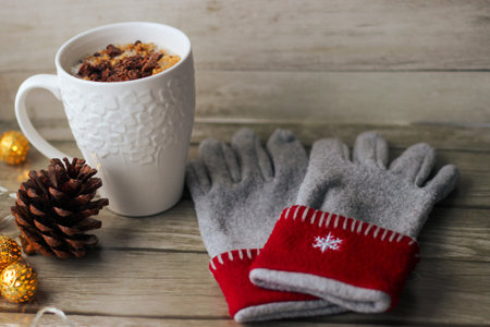 White porcelain mug filled with milk and corn flakes. Nearby are gray-red gloves with embroidered...
