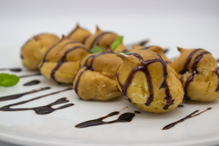 A close-up view of cream-filled profiteroles drizzled with smooth chocolate sauce. The golden pastries are accented with fresh mint leaves, making this dessert an enticing treat.の写真素材