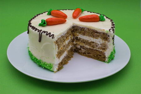 A carrot cake with cream frosting and decorative marzipan carrots, featuring a visible slice to show the layered interior. Chocolate drizzles and green accents complete the design.の写真素材