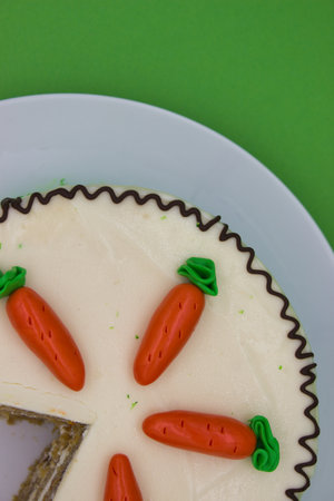 A partial view of a carrot cake topped with marzipan carrots and chocolate frosting accents. The creamy white frosting contrasts with the vibrant green and orange decorations.の写真素材