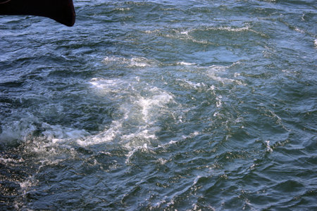 Closeup of ship stern and swirling sea water created by engine movement. Dynamic texture of waves and foam under sunlight.の写真素材