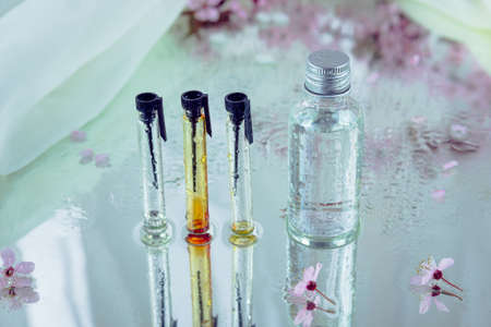 A set of three glass testers and a bottle covered with water drops and placed on a wet mirror together with blurred pink blossom. Essential oils and herbal extracts concept.の写真素材