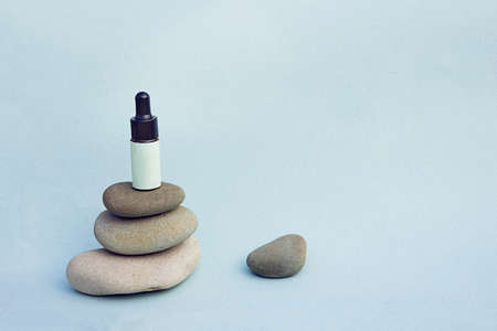 Bottle with blank spot lable and black cover with pipette on top of grey pebbles stack with one pebble on the distance with much copy space on blue background. Beauty care product bottle mockup.の写真素材