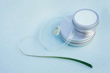 Two silver tins and a lab cup with smudge of grey paste and a long green leaf with dry edges on the side with pale blue background. Organic and eco freindly cosmetical and dental care products concept.の写真素材