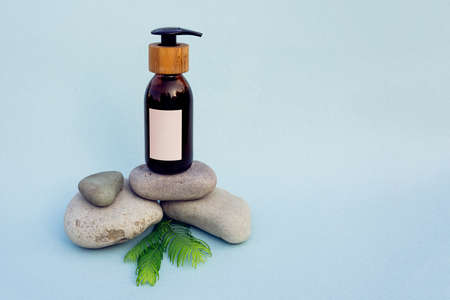 Brown glass bottle with blank spot label, black  dispencer and wooden part on top of grey pebbles with few green brunches on blue background. Care product sample with concept of natural and organic sources.の写真素材