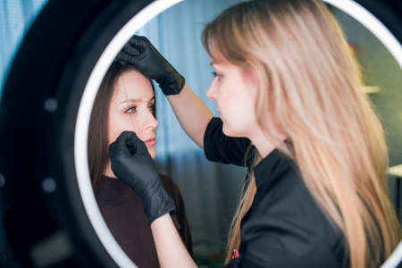 Makeup artist holds a thin coloring thread against the face of a client, marking an outline of brows, all in frame of ring light lamp. Permanent make up and brow making professional sphere.の写真素材