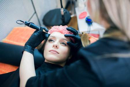 Closeup of beautiful woman's face with tatoo artist's hands holding tatoo machine upon her forehead and some blurred equipment on the background. Tattoo, permanent make up and brow making professional sphere.の写真素材
