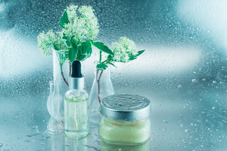 Glass flasks with flowers, a bottle of oil and big jar of cream on a silver background covered with...