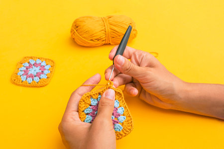 Closeup of woman's hands with a hook inserted in crochet element with the backgound of yellow table,...