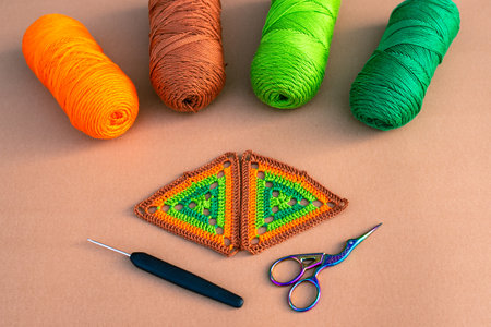 Two multicolored crochet motifs in green and orange tones, four skeins of yarn, crochet hook and small scissors on beige background. Concept of crochet supplies.の写真素材