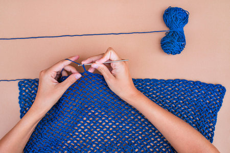 Top view of hands crocheting a blue net on beige backgorund with a blue skein of yarn above. Simple...