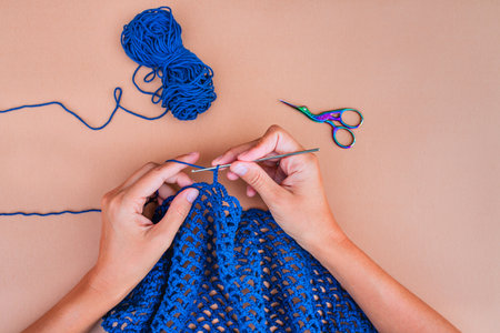 View of hands crocheting a blue net on beige backgorund with a blue skein of yarn and blue-violet scissors above. Simple and minimalistic view on crocheting.の写真素材