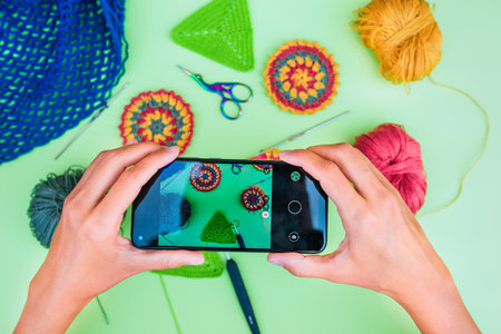 Top view on female hands making picture of layout with a black phone on light green background. Layout conteins crochet patterns, hooks and scissors.の写真素材
