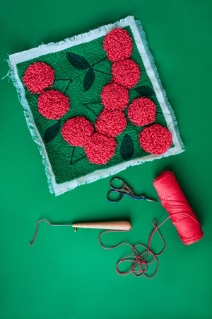 Layout of canvas with red embroidered flowers on green, punch needle, red skein of yarn, scissors on solid green background. Top view.の写真素材