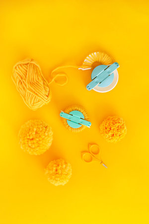 Flat lay of yellow pompoms, bulky yarn and tools for making pompoms on yellow background. Fiber arts and crafts.