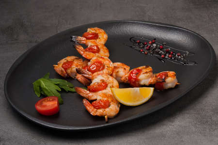 tasty grilled shrimp on a skewer with tomatoes on a dark plateの写真素材