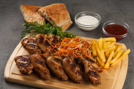 tasty grilled chicken wings with french fries, salad and sauces on a wooden boardの写真素材