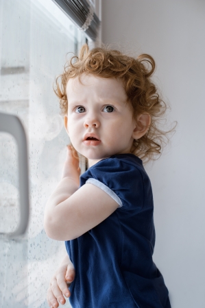 The little curly girl in a dark blue dress at a windowの写真素材