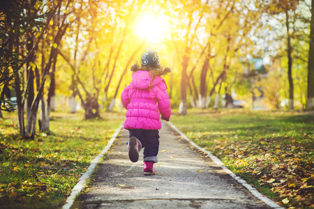 Little girl in a red jacket, running away the road in the autumn park.の写真素材