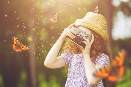 Little girl in straw hat, rustic style dress, photographing butterfly with retro photo camera in fairytale forest.の写真素材