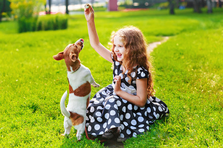 little beautiful young girl in polka dot dress, playing with her puppy jack russell in park. Happy friends leisure outdoors.の写真素材