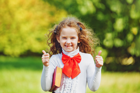 Schoolgirl in school uniform  showing thumbs up in the park. Back to school, happy childhood, successful concept.の写真素材