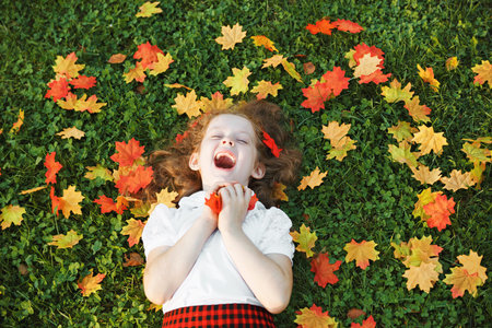 Laughing little girl lying in grass throws the autumn leaves in the air. Back to school, happy...