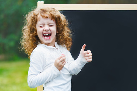 Laughing little girl showing white teeth, closed her eyes  and make gestured thumbs up, near school board.の写真素材