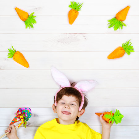 Cute child in rabbit ears with orange carrots, stuck out his tongue, lying on the white floor. Happy...