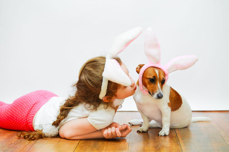 Cute girl and her friend dog with bunny ears sit on wooden floor on  light wall background.