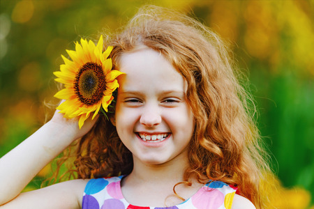 Cute little girl with sunflowers in her curly redhead hairs, enjoying nature in summer sunny day. Show white teeth. Healthcare, freedom and happy childhood concept.の写真素材