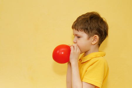 Child in yellow t-shirt inflates a red balloon on yellow background.の写真素材