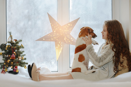 Little girl embracing her puppy dog, sitting on window and waiting Christmas.の写真素材